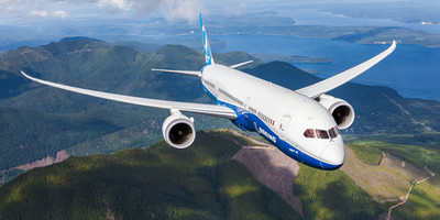 A Boeing 787 Dreamliner aircraft flying over a coastal and mountainous terrain.
